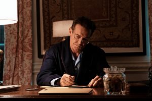 Dennis Quaid as Ronald Reagan in his film "Reagan", sitting at a desk in his pajamas and blue robe, writing something on a notepad, a look of concentration on his face.