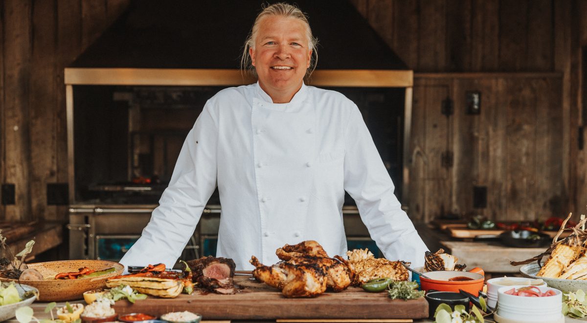 Chef Joshua Drage smiling in his kitchen with a chicken dish sitting ready on his cutting board.