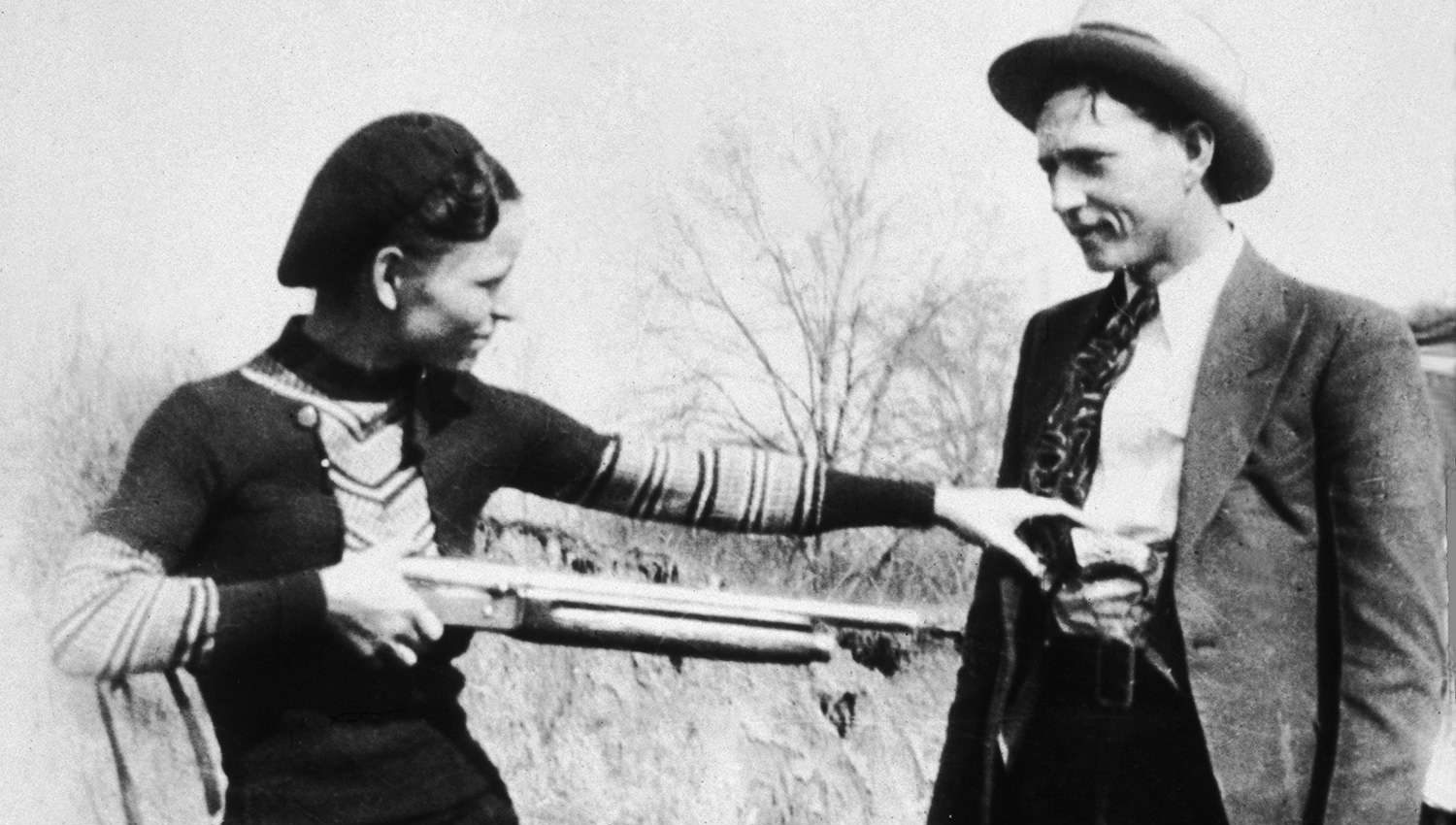 Bonnie Parker jokingly points rifle at Clyde Barrow in black and white historic photo.