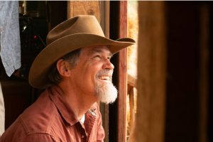 Josh Brolin smiles while looking out a barn window, wearing a brown felt cowboy hat and a rust red pearl snap shirt.