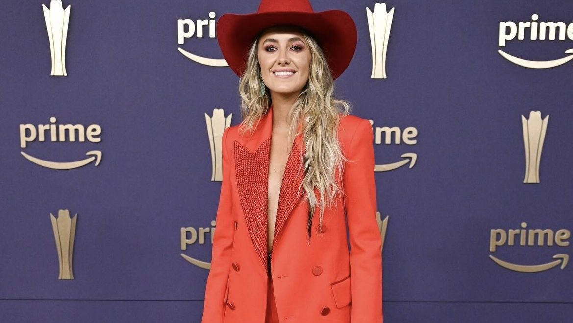 Lainey Wilson smiles on the ACM Awards 2024 red carpet wearing a red pantsuit and red cowgirl hat.