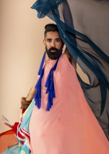 A model poses in front of a tan backdrop in a flowy colorful garment by Jason Baerg.